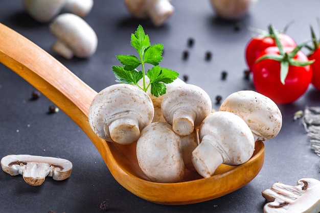 White mushrooms in wooden spoon with parsley, dill, tomatoes cherry and fresh sliced champignon on table for cooking homemade delicious dishes