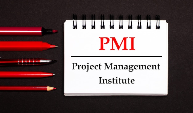 Белый блокнот с текстом PMI Project Management Institute, написанным на белом блокноте рядом с красными ручками, карандашами и маркерами на черной поверхности.
