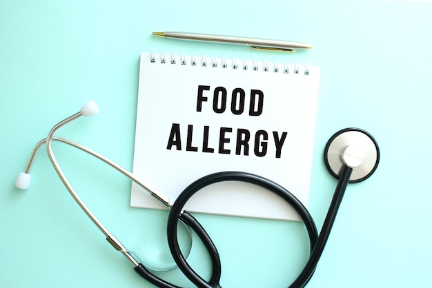 White notepad with the words FOOD ALLERGY and a stethoscope on a blue background. Medical concept