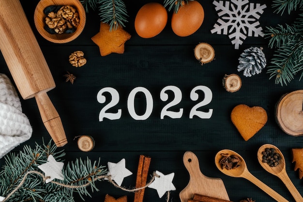 White numbers 2022 lying on black table with Christmas tree, baking accessories and ingredients. New Year 22. Top view