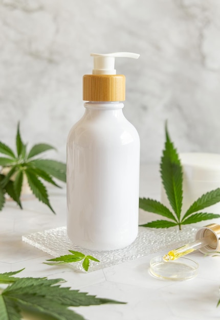 白いテーブルの上の緑の大麻の葉の近くにある白い 1 つのポンプ ボトル化粧品のモックアップ