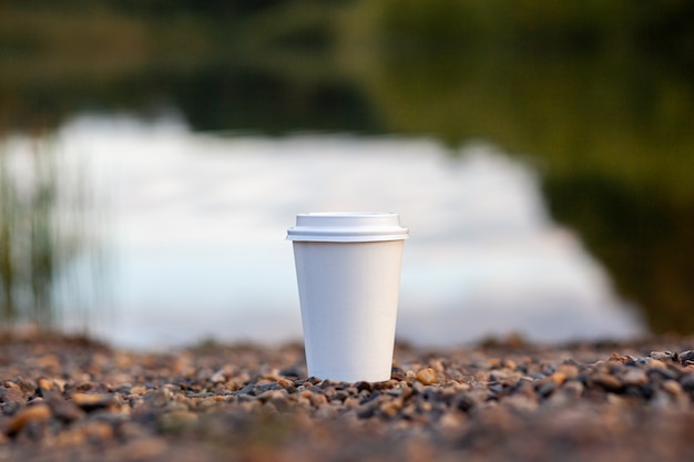 湖の岩の多い岸に白い紙コップのコーヒーまたは紅茶が立っています。小石に温かい飲み物のマグカップ。後ろの森の湖の美しい自然の背景。空きコピースペース。