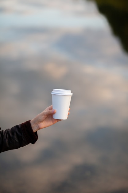 女性の手にコーヒーと白い紙コップ。街でコーヒーを飲む時間。行くコーヒー。瞬間を楽しんで、休憩してください。使い捨ての紙コップのクローズアップ。美味しい温かい飲み物。テキスト、モックアップ用の空白スペース