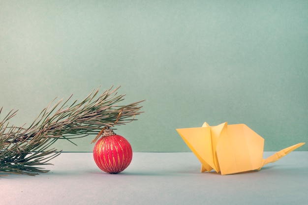 White paper mouse made using the origami technique looks at a pine branch with a red ball