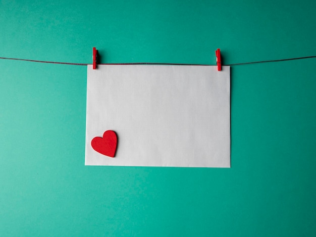 white paper that weighs on a red rope and is secured with two clothespins and has a red heart on it on a green background. a template for your project for valentine's day