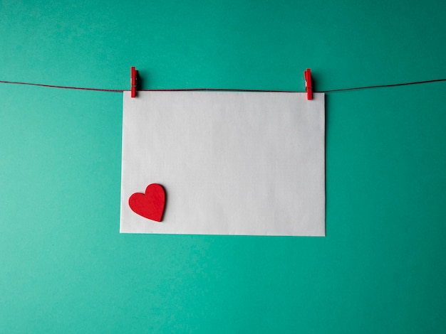 White paper that weighs on a red rope and is secured with two clothespins and has a red heart on it on a green background. A template for your project for valentine's day