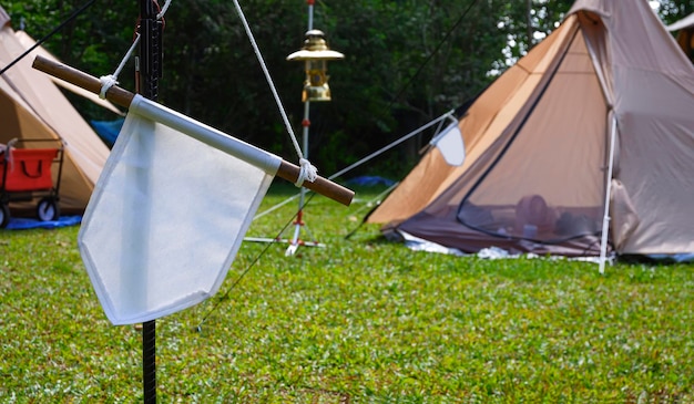 White pennant hanging on black metal pole with field tents in\
campsite area at natural parkland