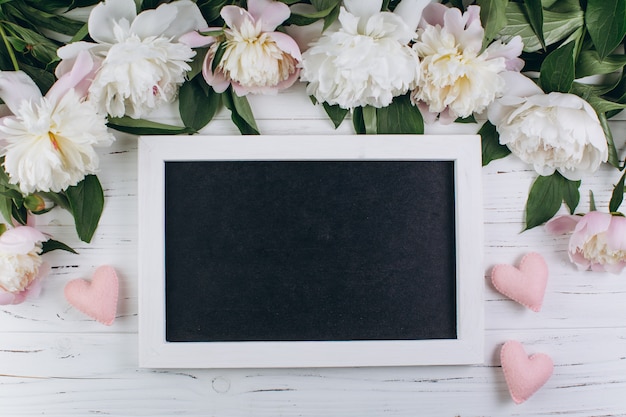 White peonies and pink hearts around blackboard on a wooden background. 