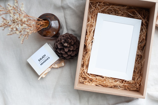 A white picture frame placed in a brown gift box surrounded by a box of small diamond rings tied with cream-colored pine cones dried flowers in a brown glass bottle.