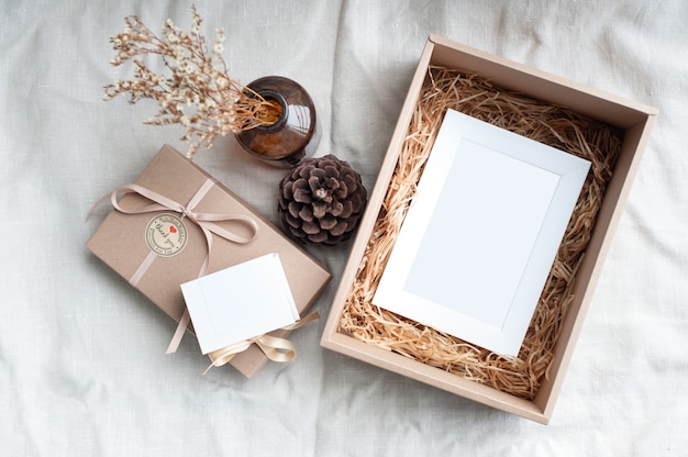 A white picture frame placed in a brown gift box surrounded by a box of small diamond rings tied with cream-colored pine cones dried flowers in a brown glass bottle. 