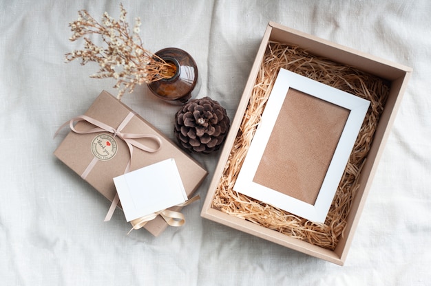 A white picture frame placed in a brown gift box surrounded by a box of small diamond rings tied with cream-colored pine cones dried flowers in a brown glass bottle. 