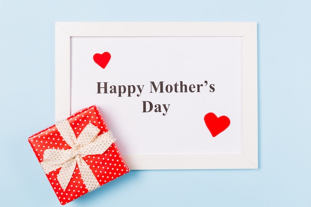 Photo white picture frame with text happy mother's day , gift box and red heart on light blue background . happy mother's day concept.