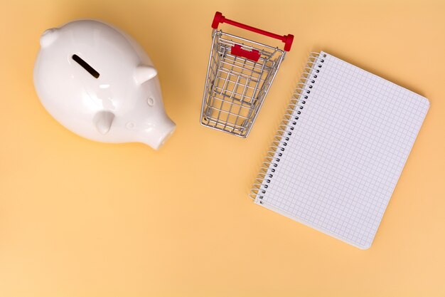 White piggy bank, shopping trolley and notebook on a beige background