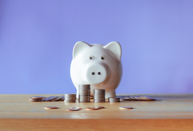 Белая копилка и стопка монет на деревянном столе с фиолетовым фоном Концепция экономии денег