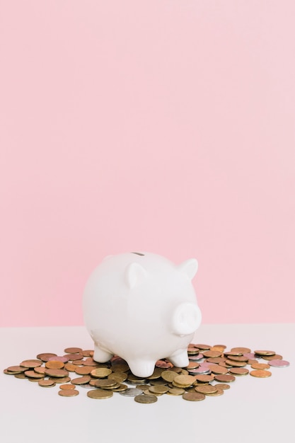 Photo white piggybank over the coins spread on white desk against pink background