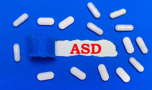 흰색 알 약은 아름 다운 파란색 배경에 거짓말. 중앙에는 ASD 자폐 스펙트럼 장애라는 비문이있는 백서가 있습니다. 의료 개념. 위에서 봅니다.
