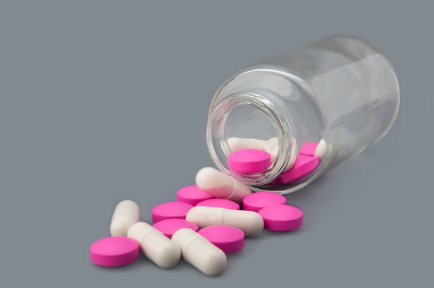 White pills and pink tablets are scattered from a glass bottle on a gray background.