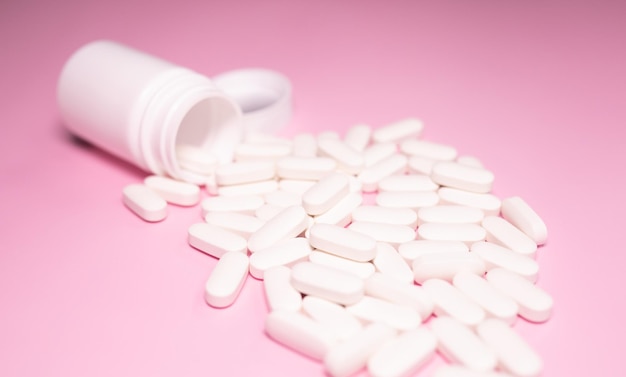 White pills spilling out of a white pill bottle on a pink background