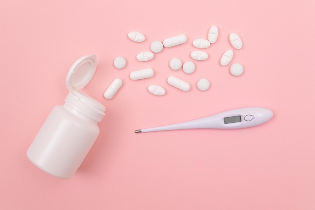 White pills or tablets with electronic thermometer on pink background