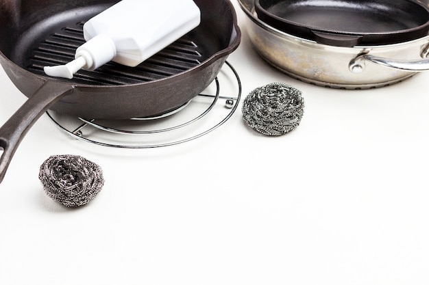 White plastic bottle in frying pan. Several frying pans and scrub buds on table. Copy space. White background. Top view