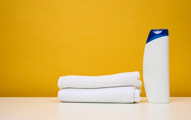 White plastic bottle for gel and shampoo and twisted white terry towels on a white table yellow background