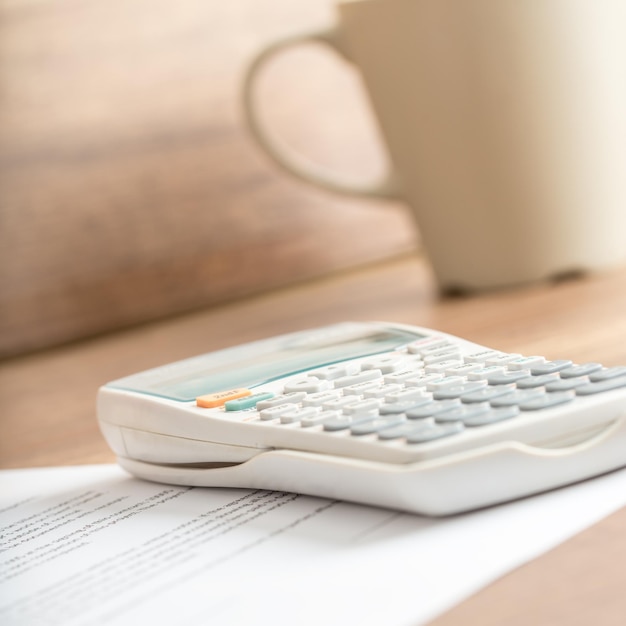 White plastic desk calculator for calculating the estimated costs written on a printed document next to a beige mug, on a wooden desk or workspace, close-up.