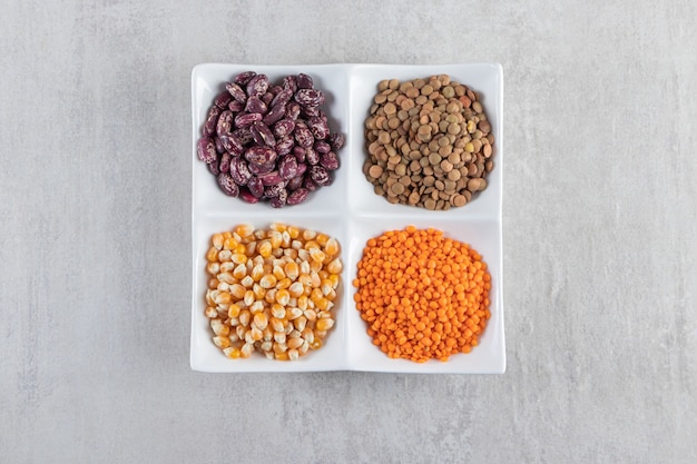 White plate full of raw lentil, corns and beans on stone background. 