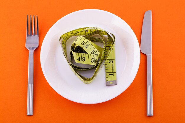 On a white plate lies a yellow measuring tape a knife with a fork on an orange surface weight loss and diet concept