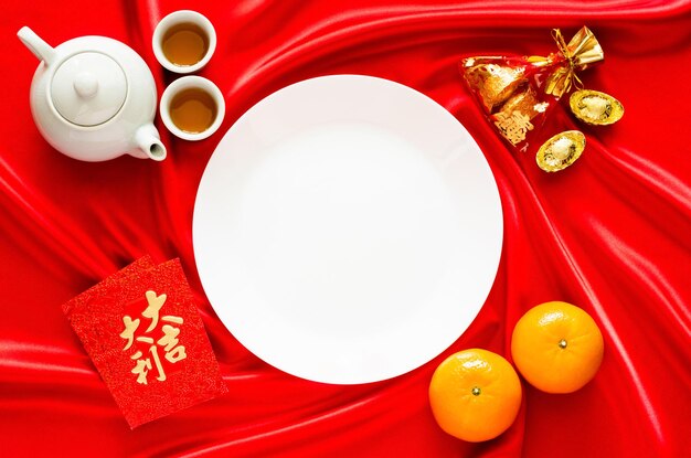 White plate on red satin cloth background with tea set, ingots,\
red bag (word means wealth), oranges and red envelope packets or\
ang bao(word means auspice) for chinese new year concept.