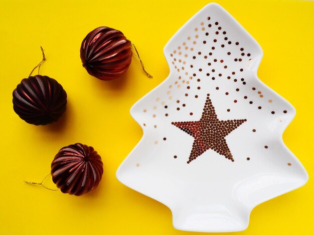 White plate in the shape of a christmas tree with gold decorations and a star in the middle yellow background new year or christmas card three beautiful shiny striped brown christmas balls