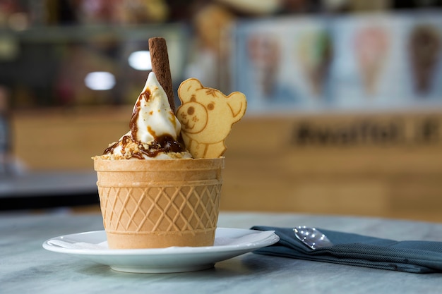 White plate with ice cream dessert in wafer cup with chocolate cookies and creative decoration topping on blurred colorful interior background.