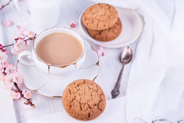 白い磁器のカップのコーヒーまたはカカオ ミルクの水差しと焼きたてのオーツ麦のクッキー イングリッシュ ブレックファーストのスティルライフ ドリンクとおやつとテーブル クロス