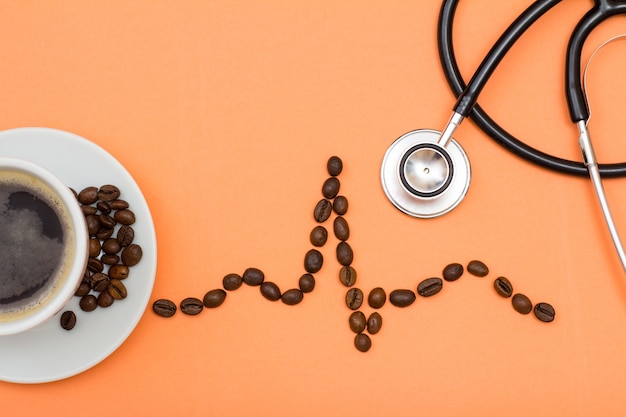 White porcelain cup of coffee on saucer with coffee beans folded in the form of a cardiogram and phonendoscope on peach colored background. Top view