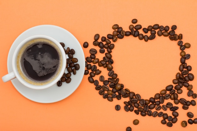 White porcelain cup of coffee on saucer with coffee beans folded in the form of a heart on peach colored background. Top view