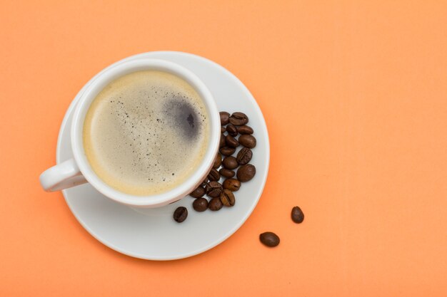 White porcelain cup of coffee on saucer with coffee beans on peach colored background. Top view