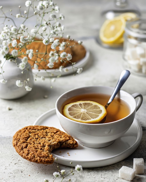 White porcelain cup with lemon tea and oatmeal cookies on textured background with flowers on the back