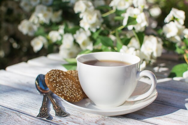 Фото Белая фарфоровая чашка с чаем и двумя овсяным печеньем с кунжутом на деревянном столе на фоне цветущего жасмина.