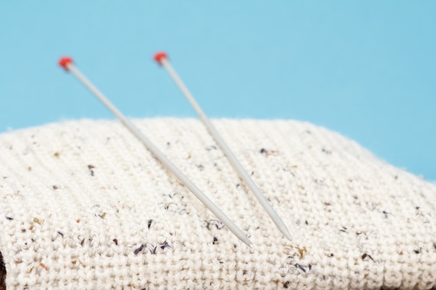 Pullover bianco e ferri da maglia in metallo su sfondo blu. concetto di maglieria. profondità di campo.