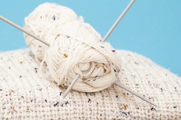White pullover and skein of yarn with metal knitting needles on a blue background. Knitting concept. Shallow depth of field.