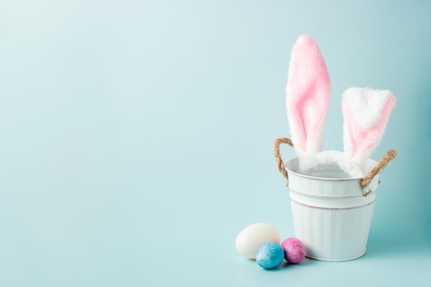 White rabbit in a white bucket and colored eggs on a blue background copy space