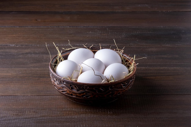White raw chicken eggs lie on hay in a burnt clay bowl
