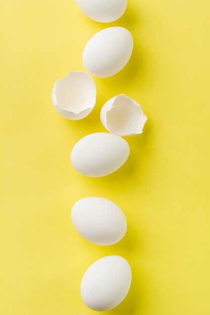 Uova di gallina crude bianche che si trovano in fila verticale con uovo rotto su sfondo giallo.