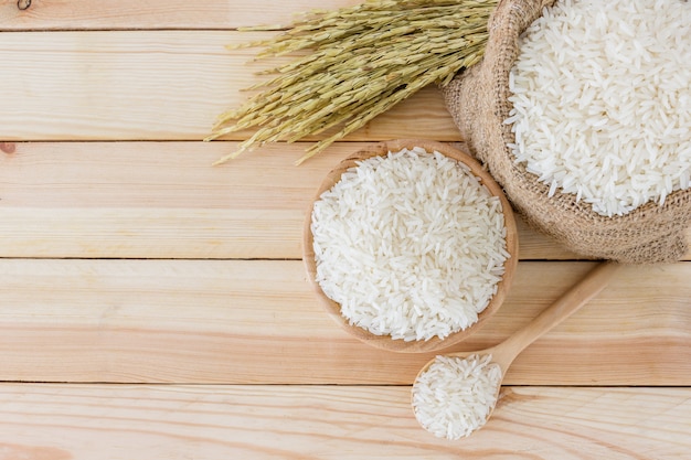 White rice in bowl and a bag, a wooden spoon and rice plant on a wooden table 