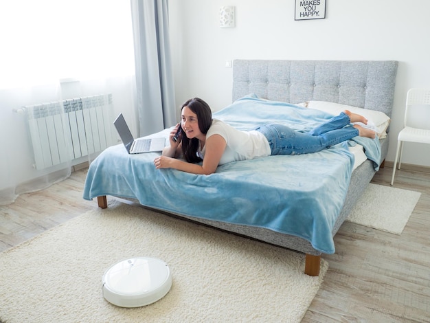 White robotic vacuum cleaner cleaning the floor while woman using laptop