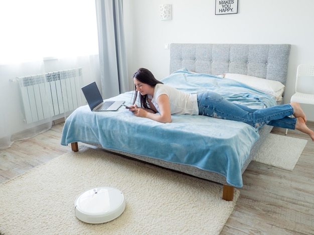 White robotic vacuum cleaner cleaning the floor while woman using laptop