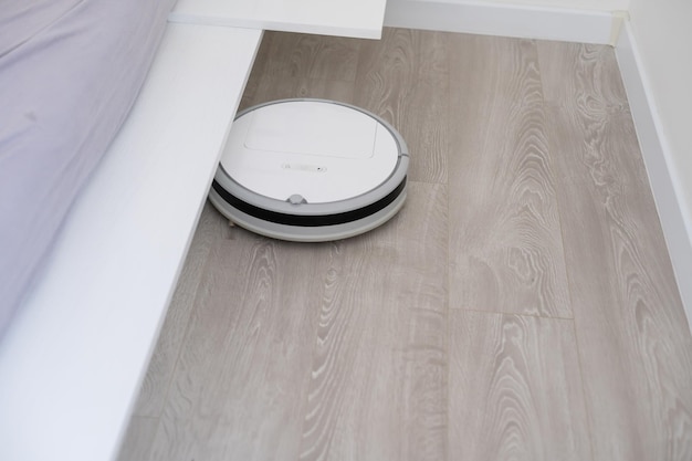 White robotic vacuum cleaner on laminate floor cleaning dust in\
living room interior. smart electronic housekeeping\
technology.