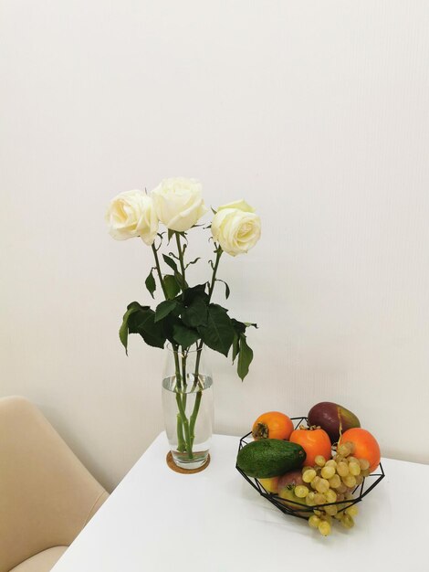 White roses in a glass vase on a white table with a fruit plate with grapes avocados persimmons