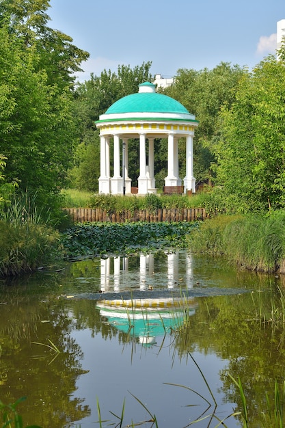 White rotunda in the park, reflection of the rotunda in the water, view of the rotunda through the trees in the park, rotunda in the summer sun