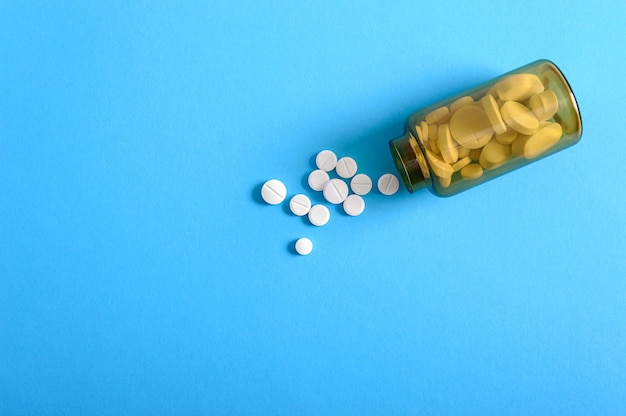 White round pills scattered from a brown bottle on a blue background. Pharmacy and healthcare theme. Medical background, copy space. Addiction treatment. Social health problems concept.