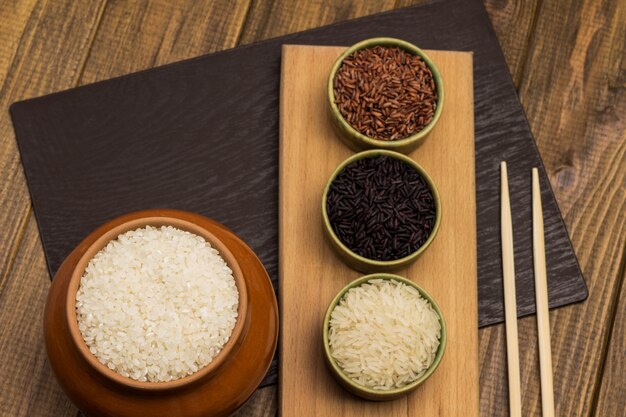 White round rice in ceramic pot.  Red and black rice in bowls. Bamboo sticks on table. Flat lay
